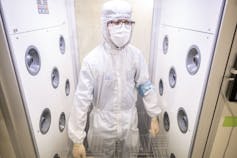 A man wears protective clothing, including gloves, a mask and a white suit, while standing in a small room with white wall panels.